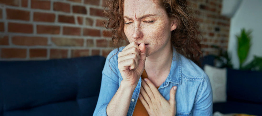 A woman sitting on a couch, coughing into her hand, representing lingering cough recovery